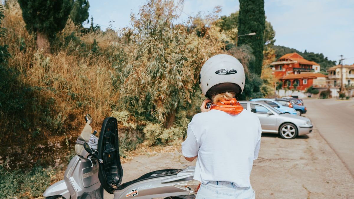 Woman wearing a helmet standing next to motor scooter in Kerkyra's rural landscape.